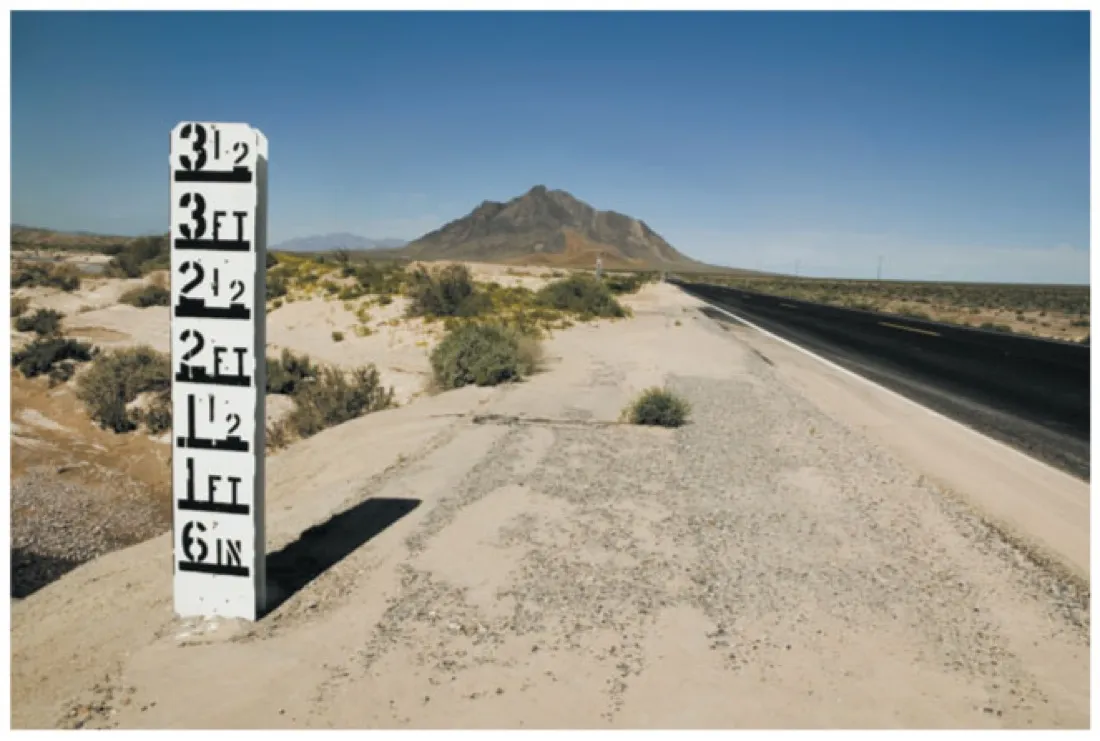 Photograph of a tall depth marker post beside a desert road, graduated from 6 inches up to 3 and a half feet