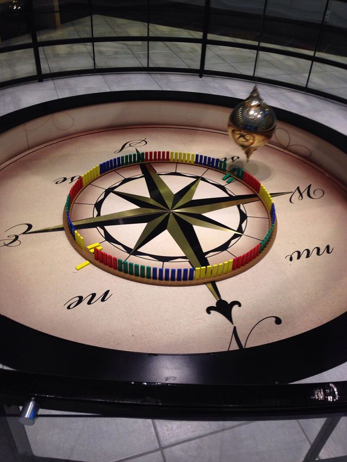 A Foucault pendulum bob swinging over a compass rose on a museum floor