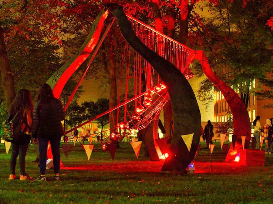 A pendulum wave machine sculpture lit red at night in a park